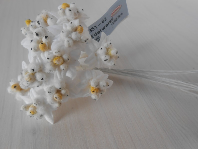 May include: A bunch of white felt edelweiss flowers with yellow centres and black accents. The flowers are attached to thin wire stems. The label reads "351 - 62 Edelweiss 1302 5cm".