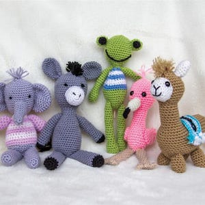 May include: A collection of six handmade crochet stuffed animals. The animals include an elephant, donkey, frog, flamingo, and llama. The elephant is lavender with pink and white striped arms. The donkey is gray. The frog is green with blue and white stripes. The flamingo is pink.