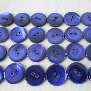 May include: A collection of twenty-four round, iridescent blue buttons. Each button has two holes and a glossy finish. The buttons are arranged in neat rows, showcasing their uniform size and color.
