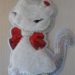 May include: White furry cat applique with a red sequin bow and a silver sequin tail. The cat has black eyes and a pink nose.