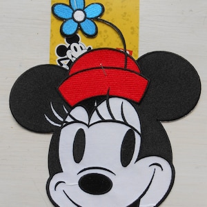 May include: Iron-on patch featuring a cartoon image of Minnie Mouse with a red hat and a blue flower on top. The patch is on a yellow background with a white border.