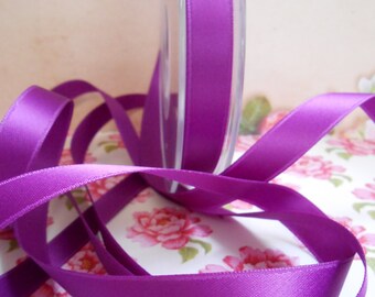 Puede incluir: Un carrete de cinta de raso morado vibrante, parcialmente desenrollada, con un brillo brillante. La cinta se muestra sobre una superficie con estampado floral con flores rosas. La cinta mide aproximadamente 2,5 cm de ancho.