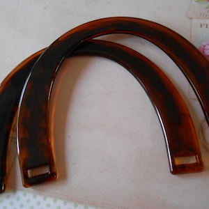 Puede incluir: Dos asas curvas para bolso, con diseño de carey. Las asas son de color marrón cálido con veteado marrón más oscuro, y tienen ranuras rectangulares en cada extremo para la fijación. Las asas son arqueadas y lisas.