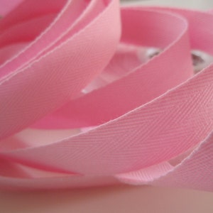 Può includere: Nastro in twill di cotone rosa con un motivo a spina di pesce. Il nastro è piegato e attorcigliato, mostrando la trama del tessuto.