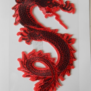 Pode incluir: Um patch de dragão bordado em vermelho e preto. O dragão está em uma pose curva, com escamas detalhadas e uma cauda fluida. O patch é provavelmente para roupas ou acessórios.