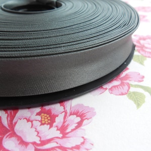 May include: A spool of dark gray satin ribbon. The ribbon is neatly wound, showing a smooth, reflective surface. The spool sits on a floral patterned fabric with pink and red flowers.