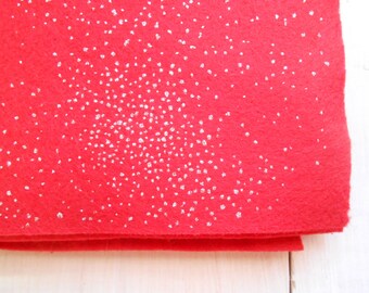 May include: A close-up of a folded red felt fabric with a scattering of silver glitter. The glitter is concentrated in the upper right corner and fades towards the bottom left. The felt has a soft, textured appearance.