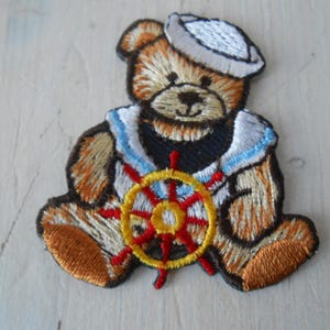 May include: Embroidered iron-on patch of a brown teddy bear wearing a sailor hat and holding a red and yellow ship's wheel.