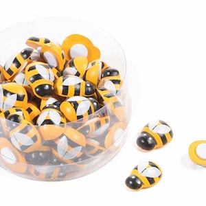 Peut inclure: Un récipient en plastique transparent rempli d'ornements en bois en forme d'abeilles. Les abeilles sont jaunes et noires avec des ailes blanches. Il y a aussi trois abeilles en dehors du récipient.