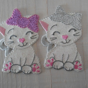 Könnte beinhalten: Zwei weiße Filzkatzen-Ornamente mit gestickten Details. Eine Katze hat eine rosa Glitzer Schleife und die andere eine silberne Glitzer Schleife.