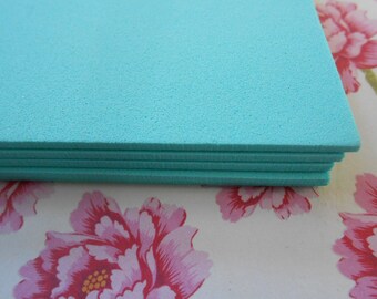 Könnte beinhalten: Ein Stapel türkisfarbener Schaumstoffplatten. Der Schaumstoff hat eine strukturierte Oberfläche und ist übereinander gestapelt. Der Hintergrund zeigt ein Blumenmuster mit rosa Blüten und grünen Blättern.