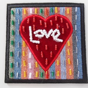Può includere: Un cuore di feltro rosso con perline bianche che compongono la parola "Love" su uno sfondo a strisce colorate. Il cuore è circondato da un bordo nero.