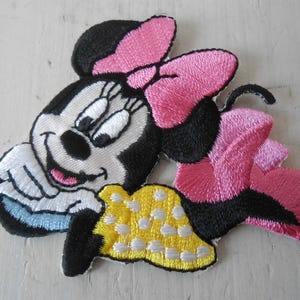 Può includere: Toppa ricamata con Minnie Mouse. La toppa mostra Minnie Mouse con testa e orecchie nere, un fiocco rosa e un vestito giallo a pois bianchi. La toppa è realizzata in tessuto e presenta un design cucito.