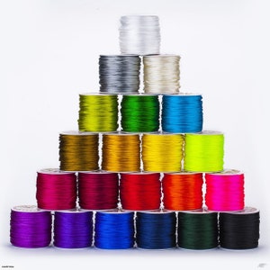 Può includere: Una piramide di rocchetti di corda colorati, disposti su uno sfondo bianco. I rocchetti presentano una varietà di colori, tra cui bianco, grigio, oro, verde, blu, rosa e nero. I cordoncini sono ben avvolti sui rocchetti.