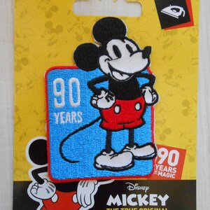 Può includere: Una toppa termoadesiva con Topolino che celebra i 90 anni. La toppa mostra Topolino con i suoi classici pantaloncini rossi e guanti bianchi, in piedi davanti a un rettangolo blu con "90 YEARS" ricamato. La confezione include il testo "MICKEY THE TRUE ORIGINAL".