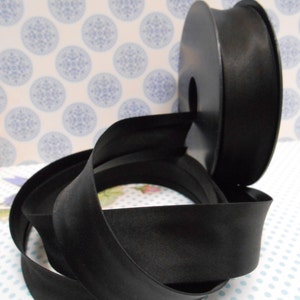 Può includere: Un rotolo di nastro di raso nero. Il nastro è avvolto attorno a un rotolo di plastica nero.