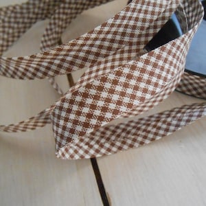 May include: Brown and white gingham ribbon with a checkered pattern. The ribbon is folded and twisted, showing the texture of the fabric.