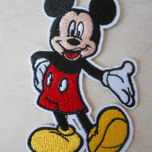 May include: Embroidered patch of Mickey Mouse in a classic pose. Mickey is shown with black ears, a black body, red shorts, yellow shoes, and white gloves. The patch has a white border.