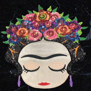 May include: A colourful embroidered patch featuring a stylised portrait of Frida Kahlo with a crown of flowers made of sequins. The patch is perfect for adding a touch of whimsy to clothing, bags, or other accessories.