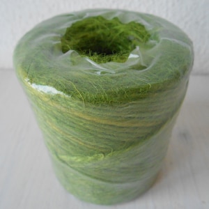 Op de afbeelding: Een cilindrische spoel van groen decoratief touw, verpakt in helder plastic. Het touw heeft een getextureerd uiterlijk met verschillende tinten groen en geel. De bovenkant van de spoel heeft een uitgehold midden, dat het binnenste materiaal onthult.