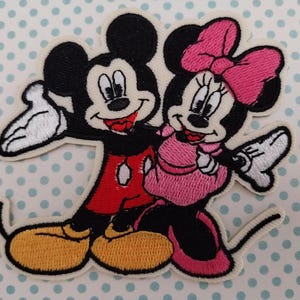 Peut inclure: Écusson brodé représentant Mickey et Minnie Mouse. Mickey porte une chemise rouge et des chaussures jaunes, tandis que Minnie porte une robe et un nœud rose. Les deux personnages sourient et saluent.