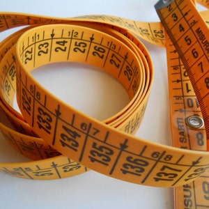 May include: A coiled orange measuring tape with black markings and numbers. The tape is marked in inches and centimeters, with the text "MISURA PER SARTI" visible. The tape is used for measuring fabric and other materials.