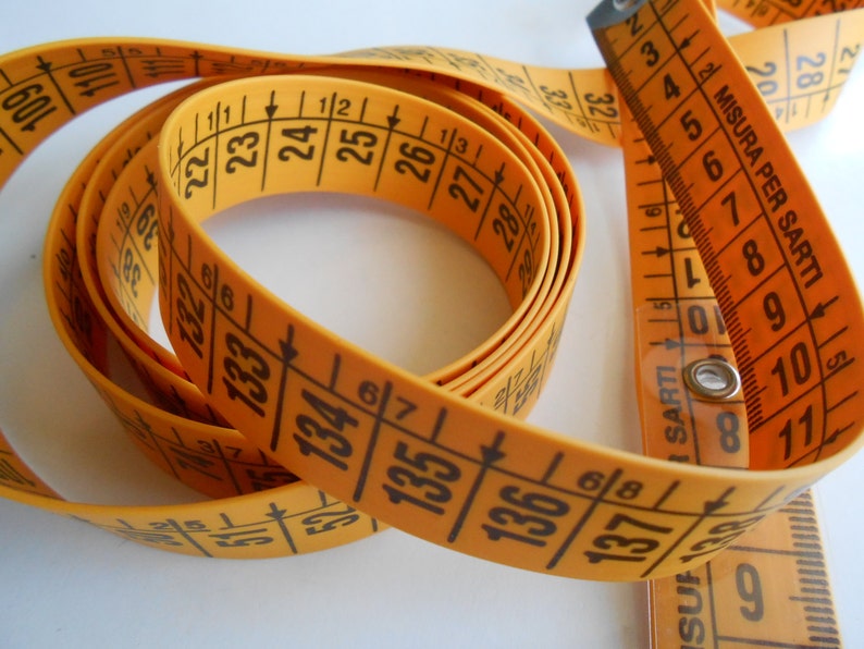 May include: A coiled orange measuring tape with black markings and numbers. The tape is marked in inches and centimeters, with the text "MISURA PER SARTI" visible. The tape is used for measuring fabric and other materials.
