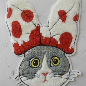 May include: A cat-shaped rug with a white face, gray and white fur, and large yellow eyes. The cat wears a large bow with red polka dots. The rug has a white border and is likely made of a soft, textured material.