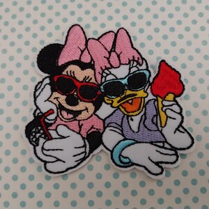 Può includere: Toppa ricamata con Minnie Mouse e Paperina con occhiali da sole. Minnie tiene in mano una bevanda con una cannuccia, mentre Paperina tiene in mano un gelato rosso. Entrambi i personaggi sorridono, con sfondo a pois azzurri.