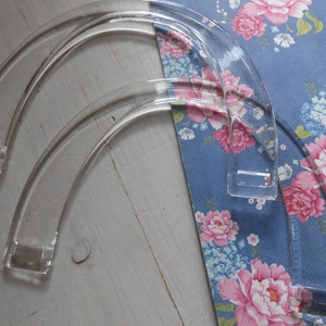 Puede incluir: Tres asas transparentes de acrílico para bolso con un diseño curvo. Las asas están colocadas sobre una tela azul con estampado floral y flores rosas y blancas. Las asas tienen puntos de fijación rectangulares.