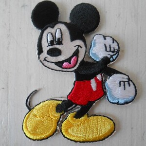 Könnte beinhalten: Gestickter Aufnäher mit Mickey Mouse in einer dynamischen Pose. Der Aufnäher zeigt Mickeys ikonische schwarze Ohren, weiße Handschuhe, gelbe Schuhe und rote Shorts. Das Design hat einen weißen Rand und eignet sich für Kleidung oder Accessoires.