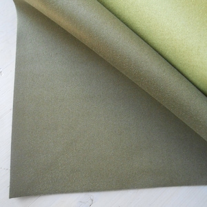 Può includere: Primo piano di un pezzo di tessuto di feltro verde piegato su se stesso. Il tessuto ha una texture sottile e un gradiente di colore da un verde più scuro a un verde più chiaro.