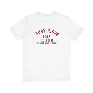 May include: A white short-sleeve t-shirt featuring red and black text on the chest. The text reads RUBY RIDGE in an arched red font, followed by 1992 in black, IDAHO in red, and the quote WE JUST WANT TO TALK in black capital letters.