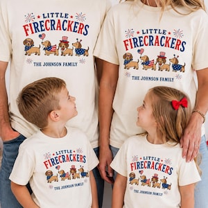 Puede incluir: Camisetas color crema con el texto "LITTLE FIRECRACKERS" en rojo y azul, con perros salchicha con atuendo patriótico. El texto "THE JOHNSON FAMILY" está debajo. Las camisetas son usadas por una familia de cuatro.