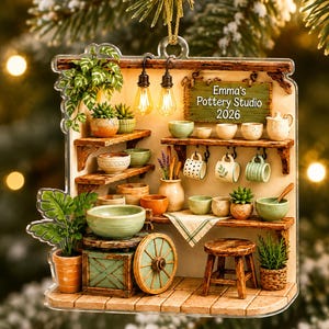May include: A flat acrylic ornament depicting a miniature pottery studio with shelves, ceramic bowls, mugs, potted plants, a wooden stool, and a pottery wheel. A sign reads Emma's Pottery Studio 2026. Two small light bulbs hang from the top frame.