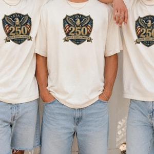 Può includere: Tre persone indossano magliette color crema e pantaloncini di jeans blu chiaro. Le magliette presentano una grafica con un'aquila, il testo "USA 250 UNITED STATES OF AMERICA 1776-2026" in un design a scudo. L'immagine è ben illuminata e l'abbigliamento sembra casual.