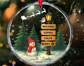 May include: A round Christmas ornament with a winter scene featuring a snowman, pine trees, and a street lamp. The ornament has the names Michael, Sophia, Chloe, and Buddy. The text "Smith Family 2025" is also visible.