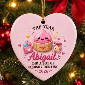 May include: A heart-shaped pink ceramic ornament featuring a cartoon pink dumpling in a bamboo steamer, a pink shopping bag with two small creatures, and a jar with a bear. Text reads: THE YEAR Abigail DID A LOT OF SQUISHY HUNTING 2026.