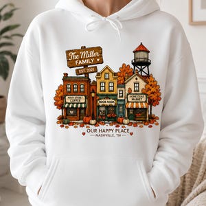Peut inclure: Un sweat à capuche blanc avec un motif coloré représentant une rue de petite ville avec des arbres d'automne, des citrouilles et un château d'eau. Le texte indique The Miller FAMILY EST. 2021, MAIN STREET COFFEE, BOOK NOOK, HOMESTEAD MARKET, OUR HAPPY PLACE, NASHVILLE, TN.