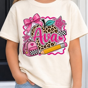 以下が含まれることがあります： クリーム色のTシャツ。ヒョウ柄のリンゴ、ピンクのリボン、チェック柄のポケットが付いたピンクのバックパック、黄色い鉛筆、2つのピンクのグリッタースターが描かれたグラフィックデザイン。リンゴにはピンク色で「Ava」という名前がプリントされています。