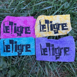 Peut inclure: Quatre écussons rectangulaires en tissu rose, jaune, bleu et violet, chacun portant le texte noir "letigre" imprimé dans une police grasse, carrée et vieillie. Les écussons ont des bords bruts et irréguliers ainsi qu'un aspect texturé.
