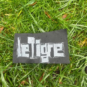 May include: A black rectangular patch with the text "le tigre" in a bold, silver, blocky font. The patch is set against a backdrop of green grass and fallen leaves.