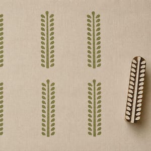 May include: A rectangular wooden printing block with a carved leaf stem pattern, positioned next to a beige fabric surface featuring six identical olive green leaf stem prints arranged in two rows of three, showcasing the stamp design.