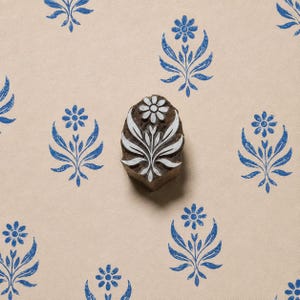 Op de afbeelding: Een donkerbruine houten blokstempel met een uitgesneden bloemmotief met een centrale bloem en symmetrische bladeren. De stempel ligt op een lichtbruine ondergrond die is versierd met meerdere blauwe inkt-afdrukken van hetzelfde bloemmotief.