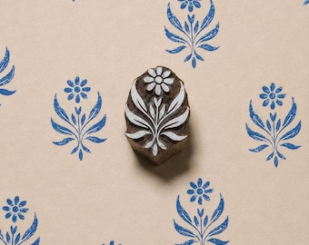 May include: A dark brown wooden block stamp featuring a carved floral motif with a central flower and symmetrical leaves. The stamp is positioned over a light tan surface printed with multiple blue ink impressions of the same floral design.
