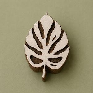 Puede incluir: Un sello de madera para estampar con forma de hoja de monstera. El sello presenta una base de madera natural de color marrón oscuro con una superficie elevada, plana y de color claro que forma el intrincado patrón de hoja perforada.