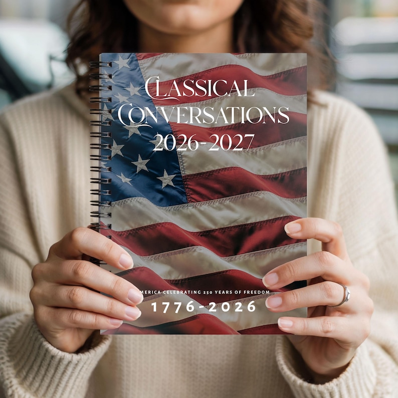 May include: A spiral-bound notebook with the American flag design. The cover features the text "Classical Conversations 2026-2027" and "America Celebrating 250 Years of Freedom 1776-2026". The notebook is held in front of a person.