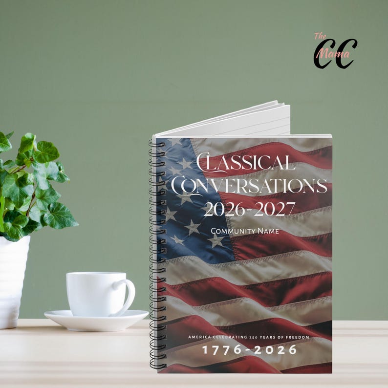 May include: A spiral-bound notebook with a cover featuring the American flag. The text on the cover reads "CLASSICAL CONVERSATIONS 2026-2027" and "AMERICA CELEBRATING 250 YEARS OF FREEDOM 1776-2026". A white coffee cup and plant are in the background.
