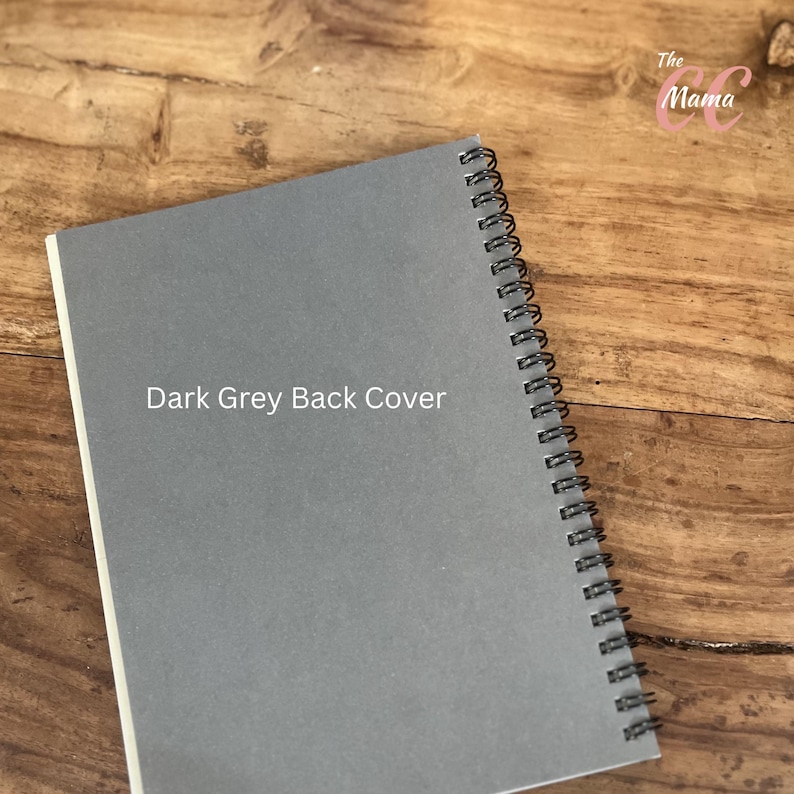 May include: A dark grey spiral-bound notebook with the text "Dark Grey Back Cover" printed on the front. The notebook is open, revealing white pages. The spiral binding is black. The notebook is on a wooden surface.