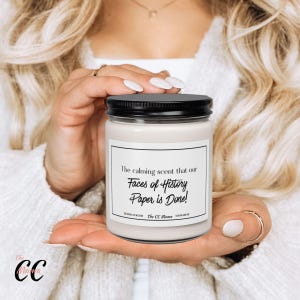 May include: A clear glass candle jar with a black lid and a white label. The label features the text: The calming scent that our Faces of History Paper is Done! The CC Mama. The candle contains a light-colored wax and is held in a neutral position.